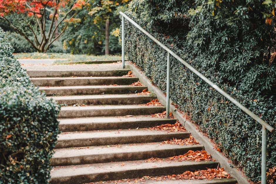 A concrete staircase outdoors, leading up through a garden area with fallen orange and brown autumn leaves scattered across each step. On the right side, a metal handrail runs parallel to the steps, s